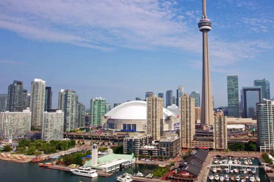 Toronto skyline featuring the CN Tower and Rogers Centre, with waterfront buildings and boats in the foreground under a partly cloudy sky.