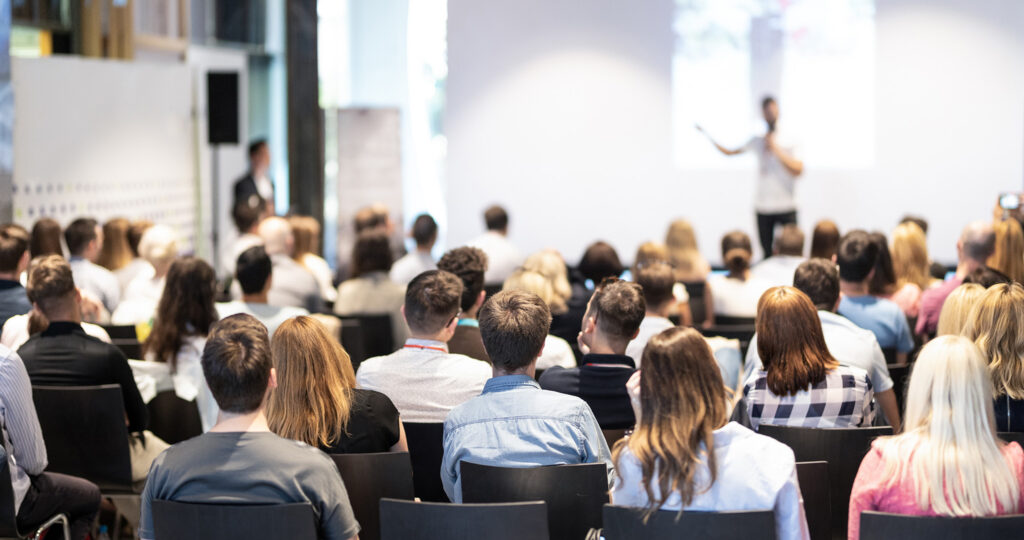 A group of people seated in a conference room listens to a speaker presenting slides at the front of the room.