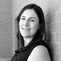 A woman with straight dark hair, wearing a sleeveless top, smiles while leaning against a brick wall. The photo is in black and white.