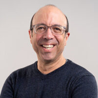 A middle-aged man with glasses and a receding hairline smiles at the camera, wearing a dark sweater and standing against a plain light background.