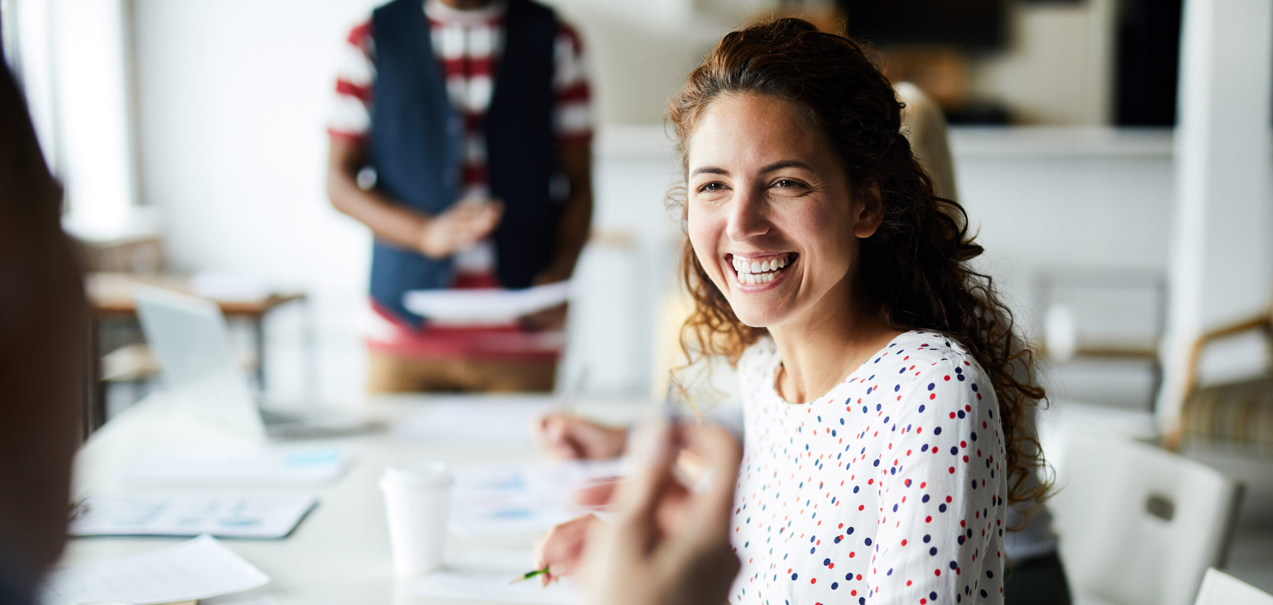 A woman smiling at a desk during a meeting, with another person blurred in the background holding papers.