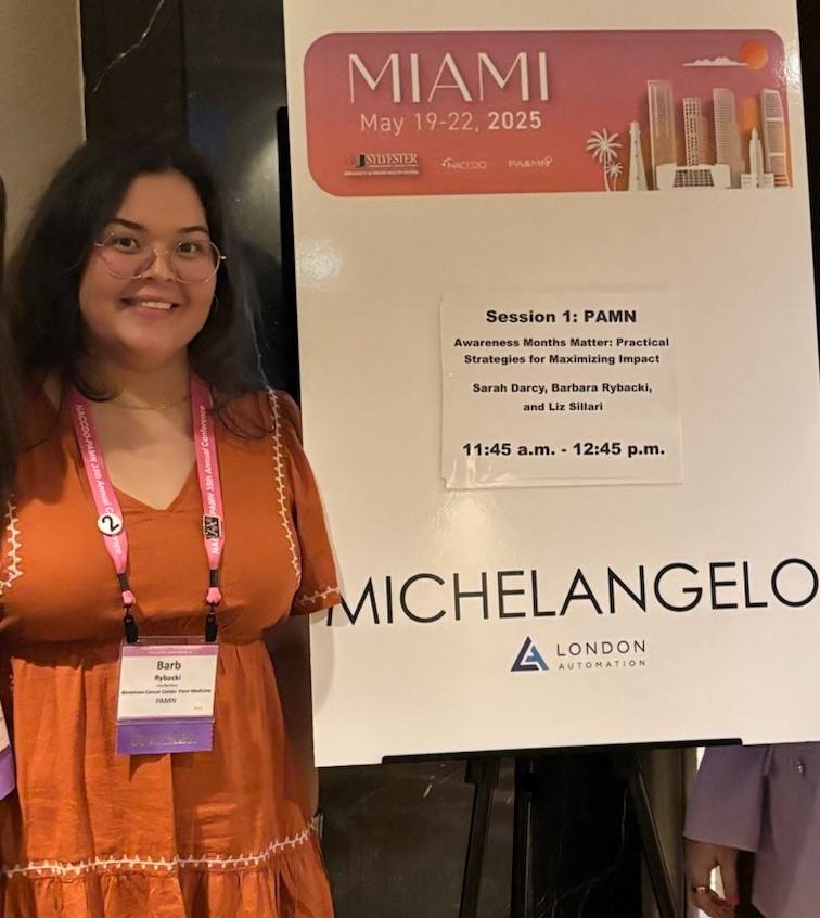 A woman wearing glasses and a pink dress stands next to a conference sign for a session titled "Awareness Months Matter" at an event in Miami scheduled for May 19-22, 2025.