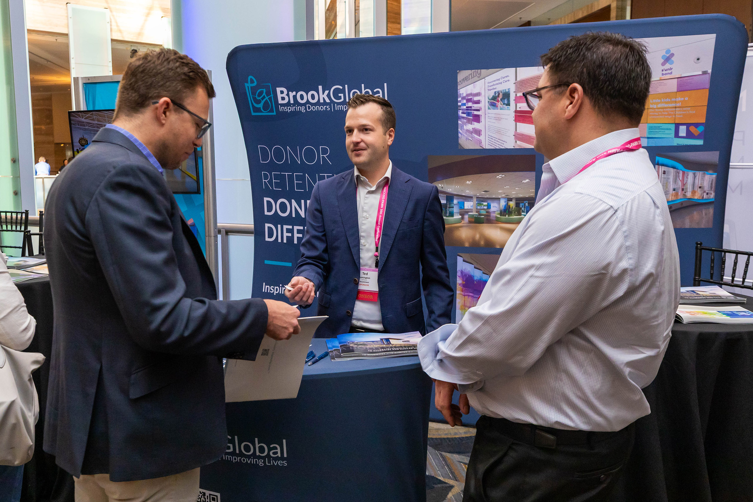 Three men in business attire talk at a BrookGlobal booth during a conference, with promotional materials and display banners visible behind them.