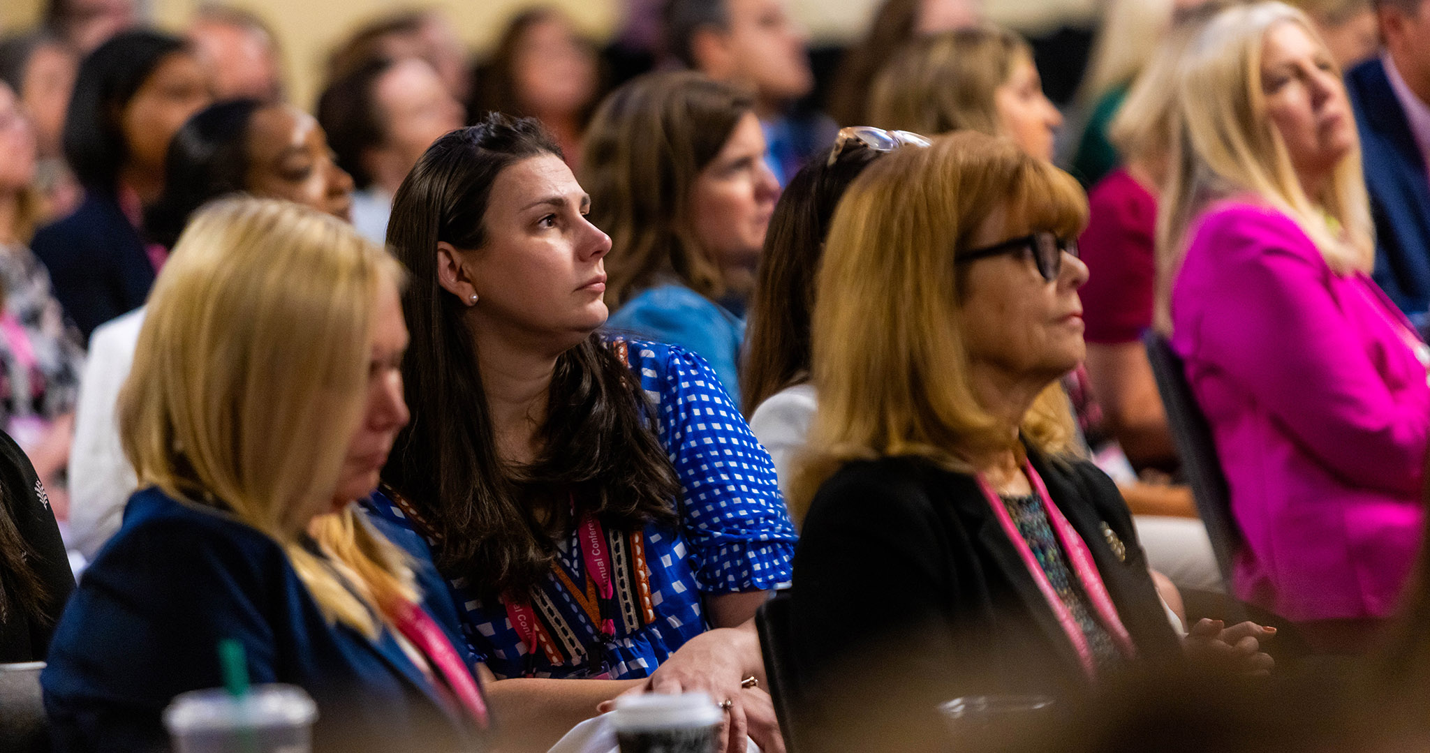 A group of people sit closely together in an audience, attentively listening to a presentation or speaker at an indoor event.