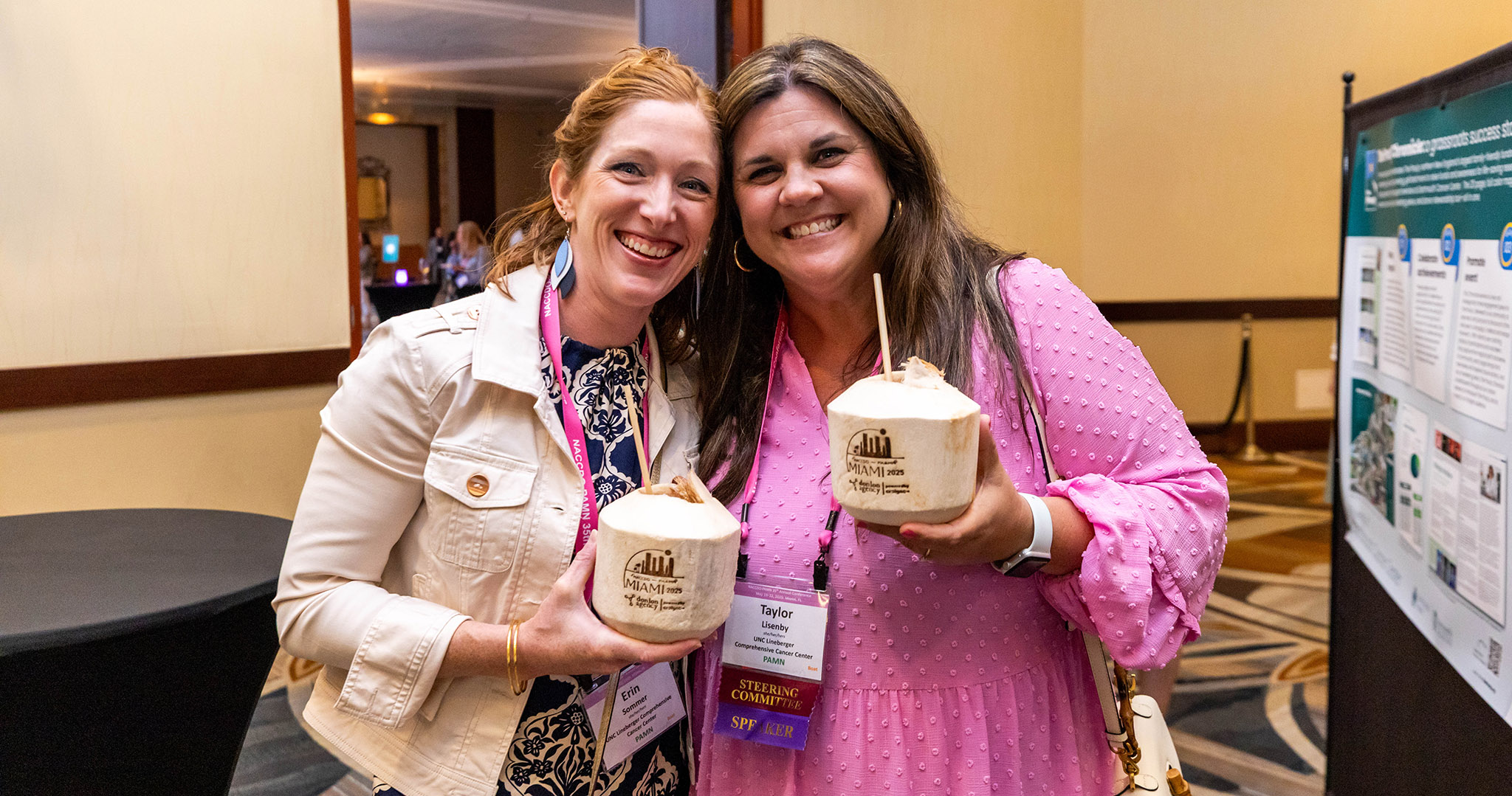 Two women stand indoors, smiling and holding coconuts with straws. Both wear name badges and conference lanyards, with a research poster visible in the background.